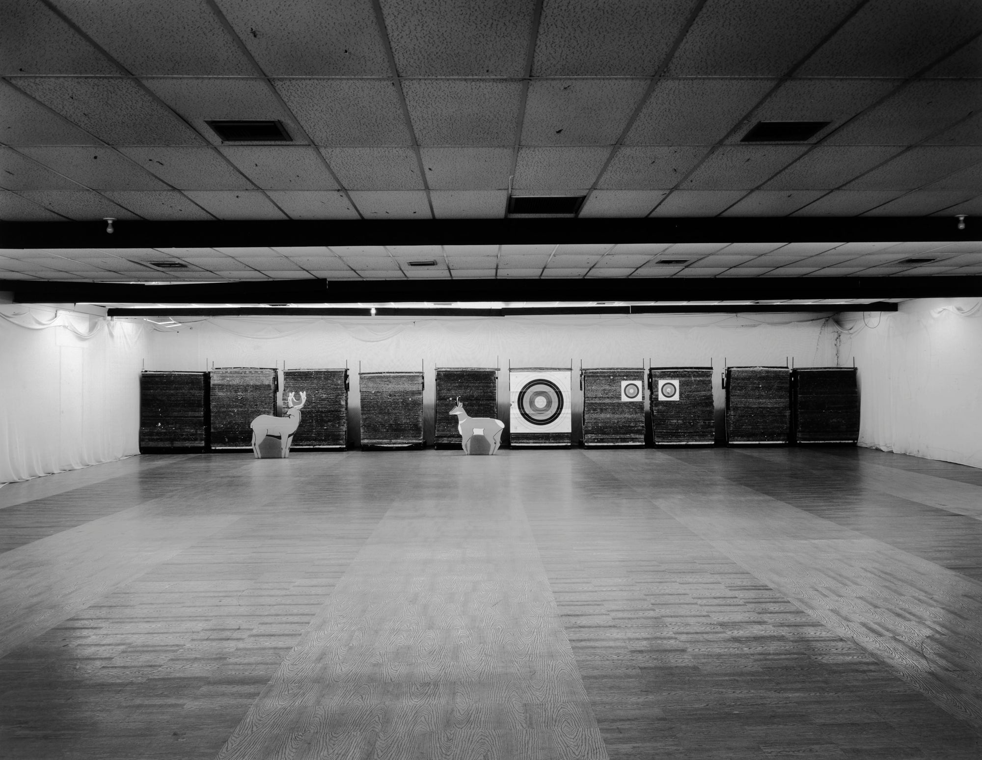 A black and white photograph of an indoor archery range, showing two deer targets and a bullseye target set against a wall of dark wooden stands, with a large open floor and ceiling visible.