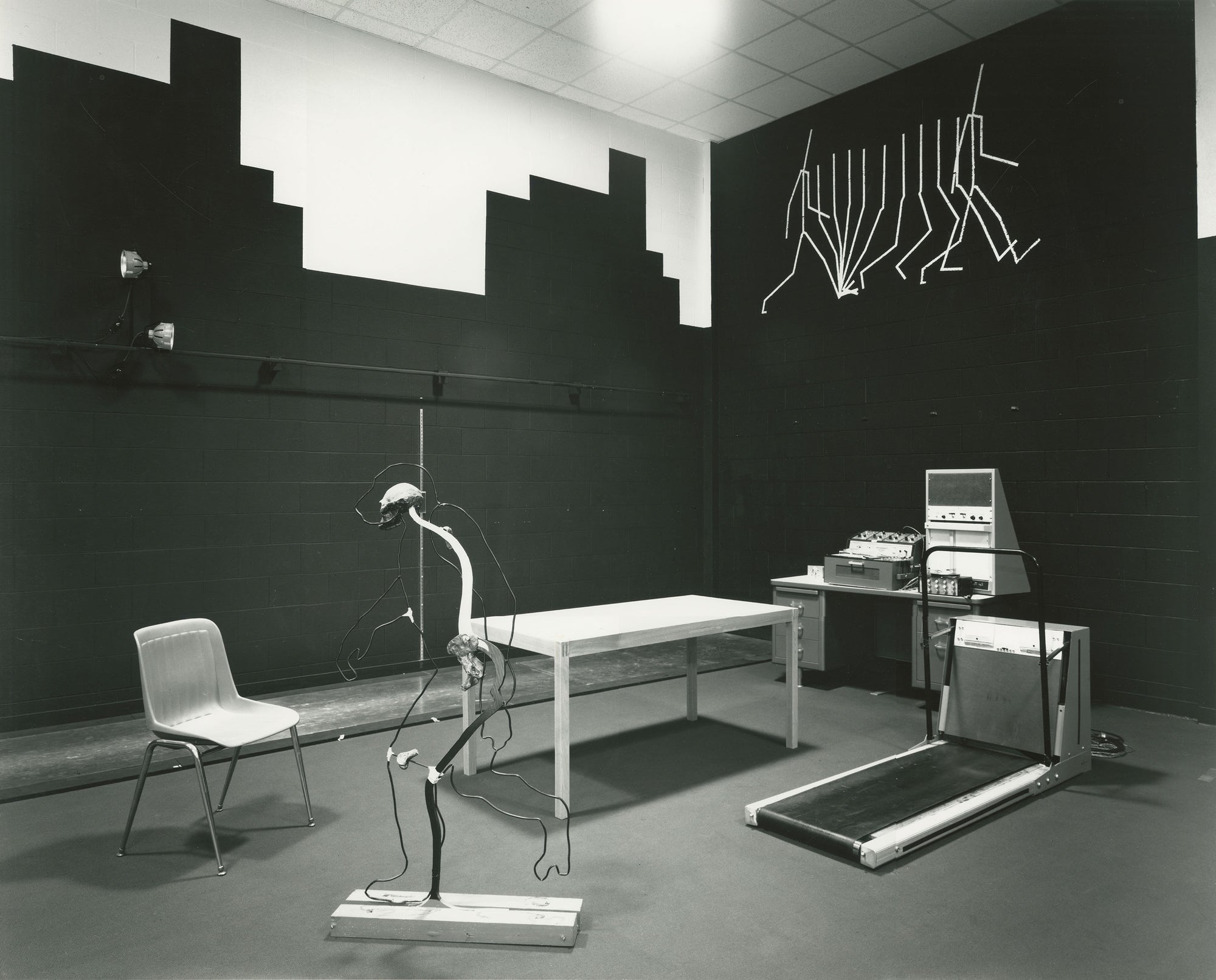 A black-and-white interior scene showing a rehabilitation or therapy room with a treadmill, a standing exercise device, a chair, a table, and wall-mounted line drawings on dark walls.