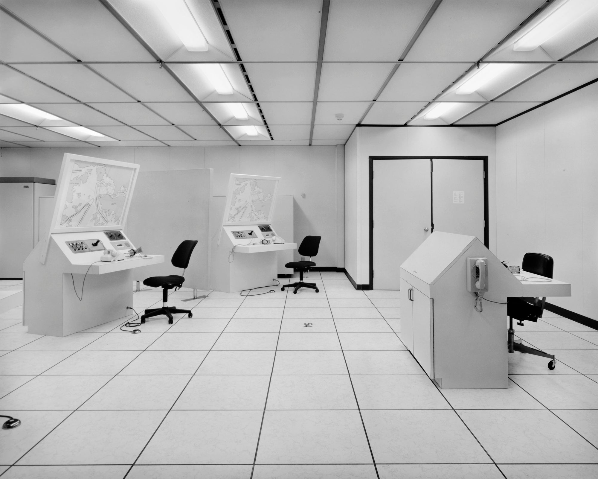 A black and white photograph of a vintage computer mainframe room with multiple terminal consoles, office chairs, a wall-mounted telephone, and a tiled floor and ceiling.