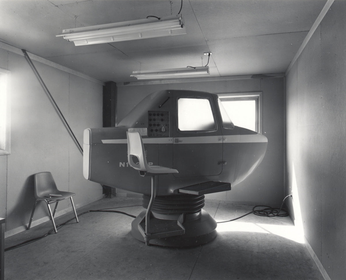 A black and white photograph of a large flight simulator cockpit or motion platform mounted in a room with fluorescent lights, walls, and a chair visible.