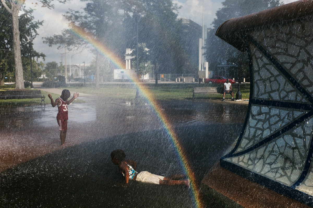 A children playing in a wet park fountain with a large rainbow arching through the spray. One child stands in the water wearing a red jersey, another lies on the wet ground in white shorts, and a mosaic-covered structure is visible on the right.