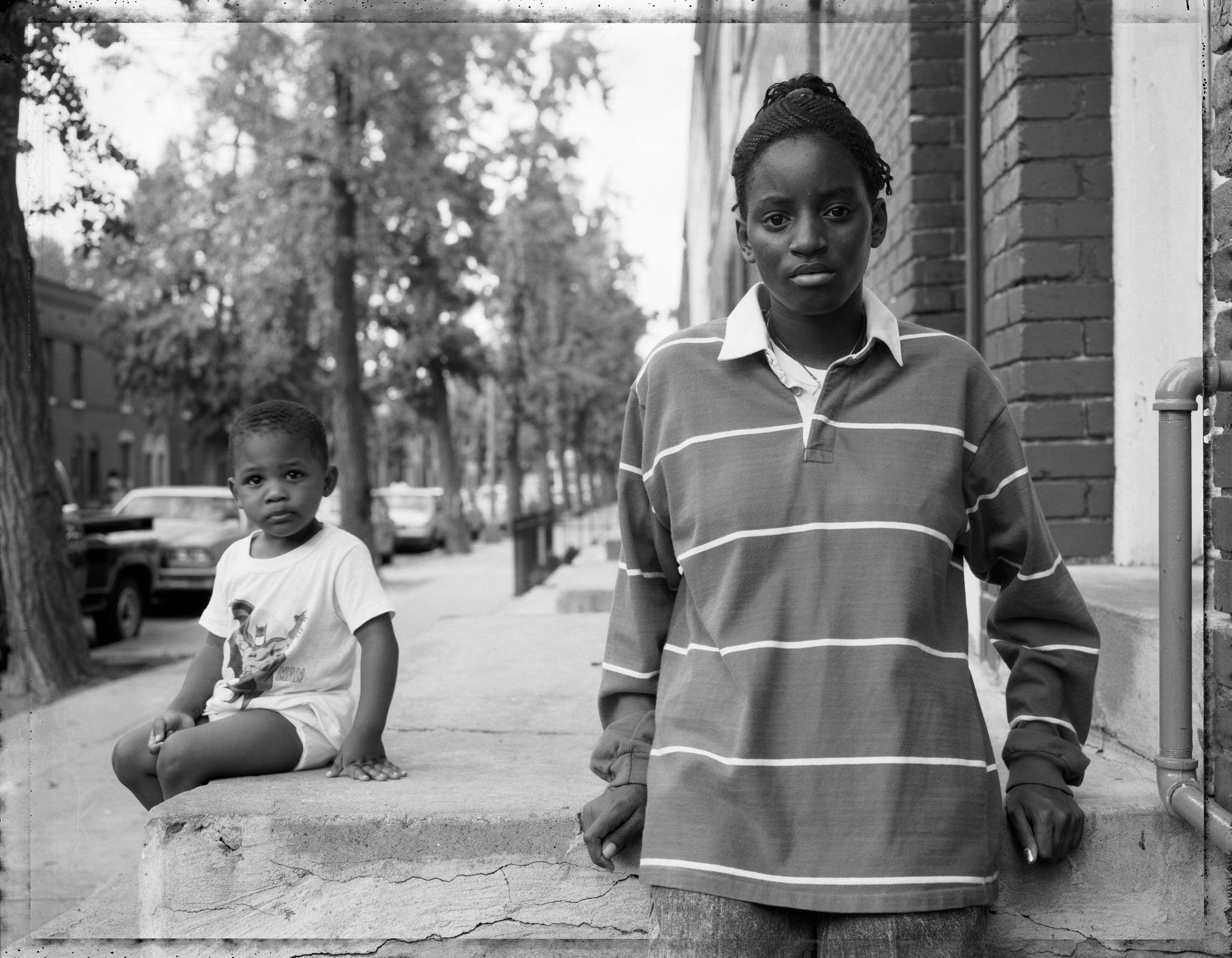 A black and white street photograph showing a woman standing on a sidewalk in front of a brick building, with a young child sitting on a concrete ledge in the background.