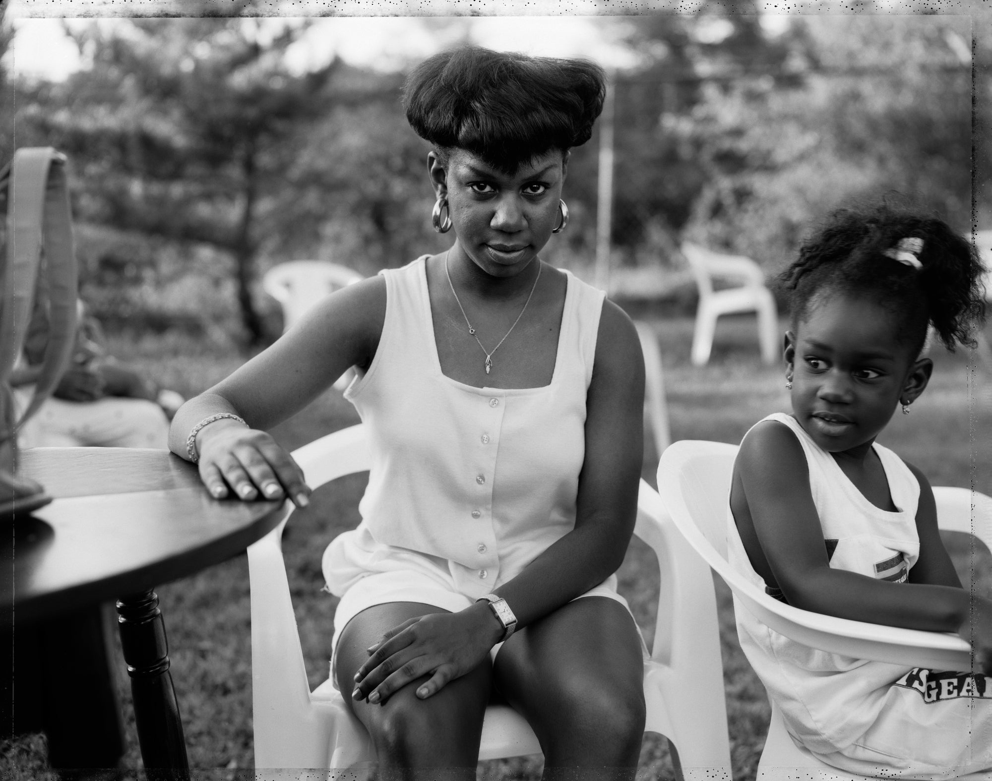 Black and white photograph of a woman and a young child sitting outdoors on white plastic chairs, with trees and patio furniture blurred in the background.