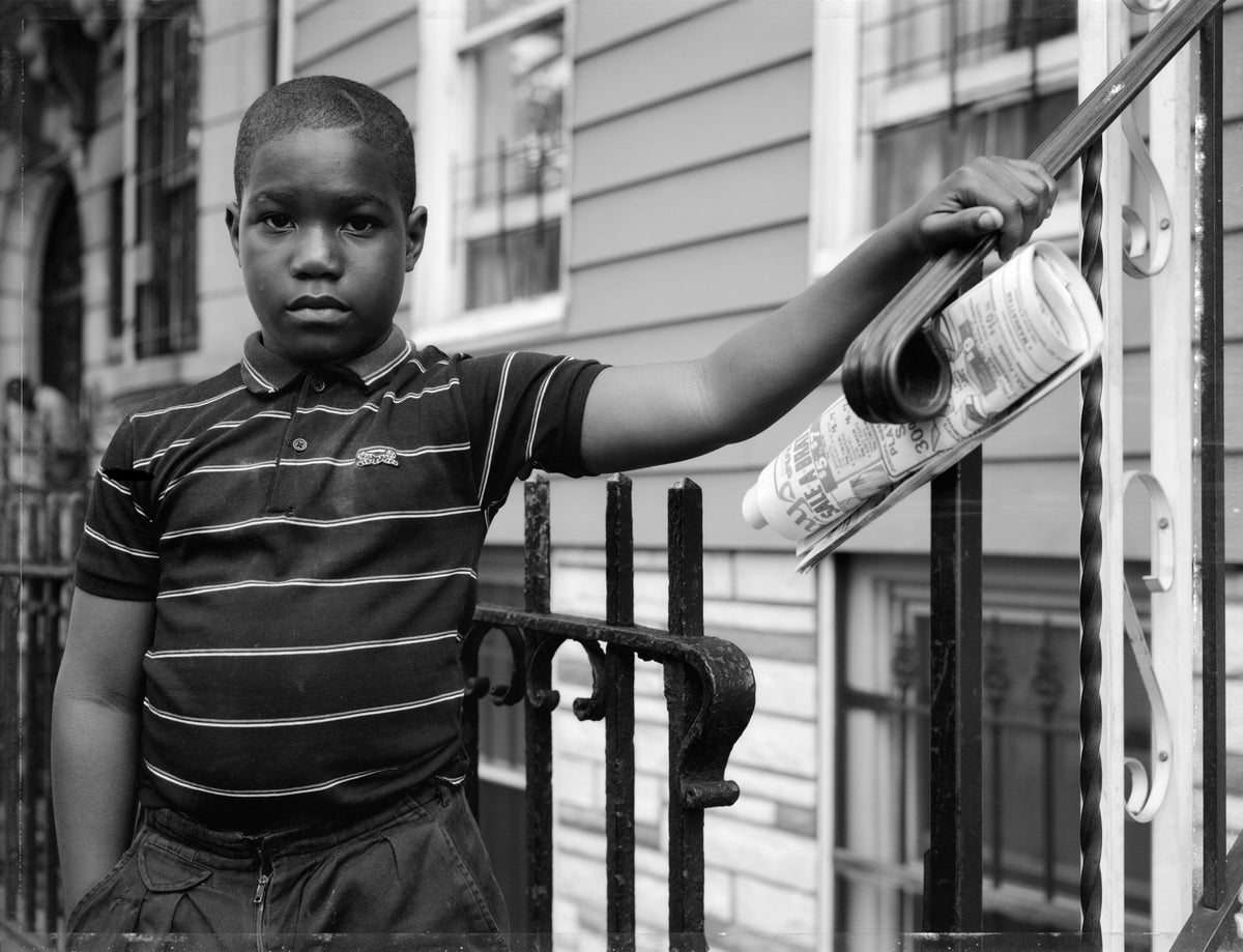 A black and white photograph of a young boy standing on a porch, holding a rolled-up newspaper against a metal railing. He is wearing a striped polo shirt and shorts.