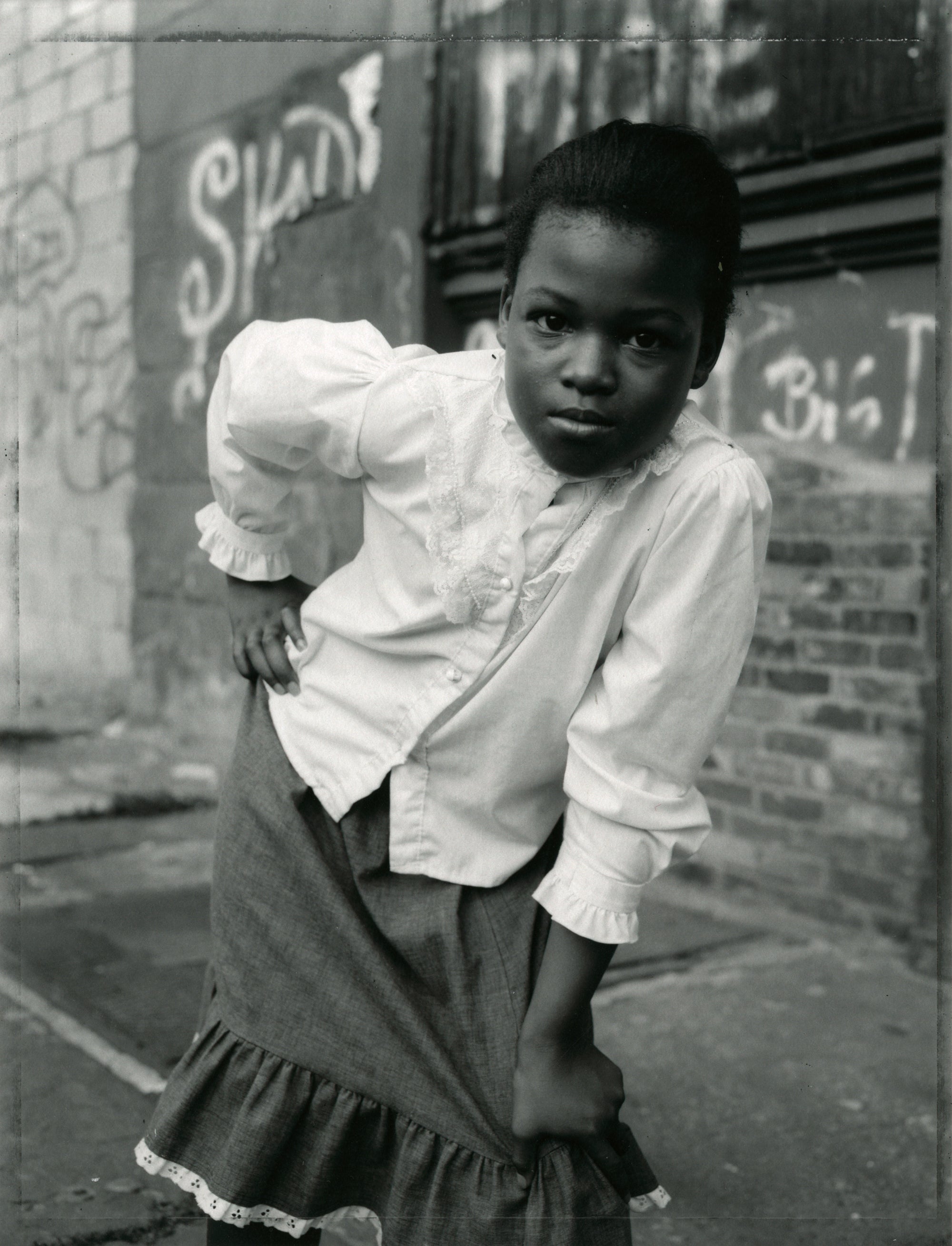 A black and white portrait photograph of a young girl standing outdoors against a graffiti-tagged brick wall, wearing a white blouse and a dark skirt.