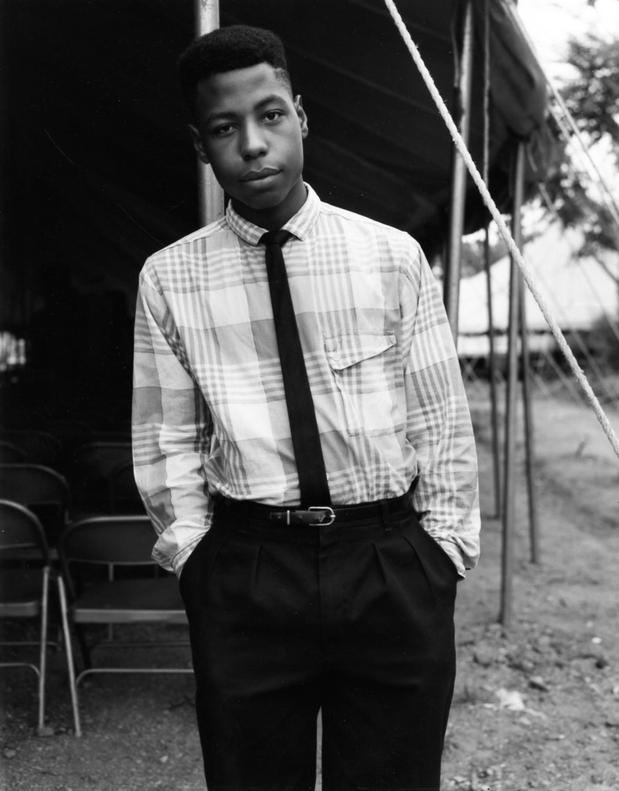 A black and white portrait of a young man standing outdoors in front of a tent, wearing a light plaid button-up shirt, a dark tie, and dark trousers.