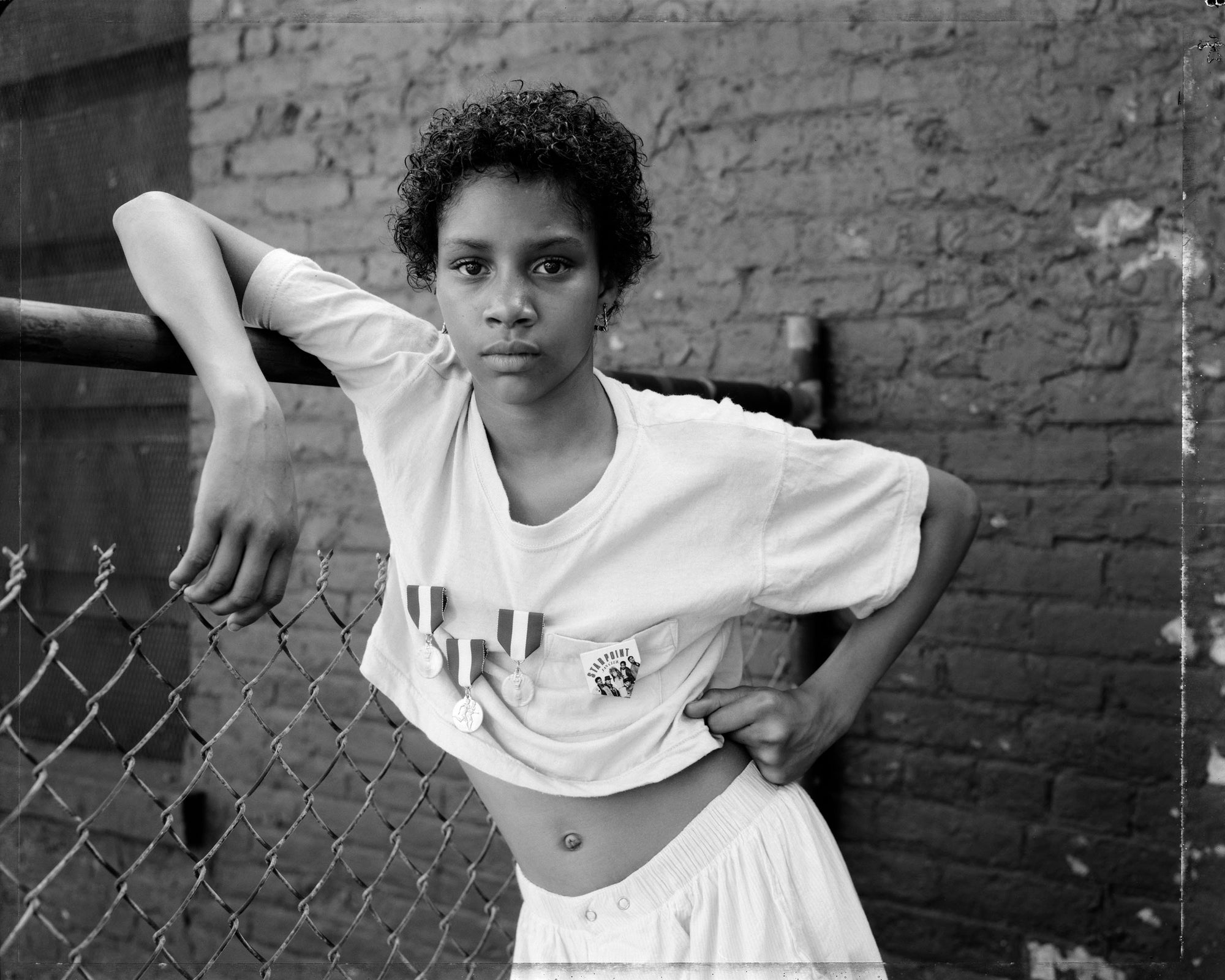 A black and white portrait of a young person with curly hair leaning against a chain-link fence and brick wall, wearing a cropped white T-shirt and light shorts.