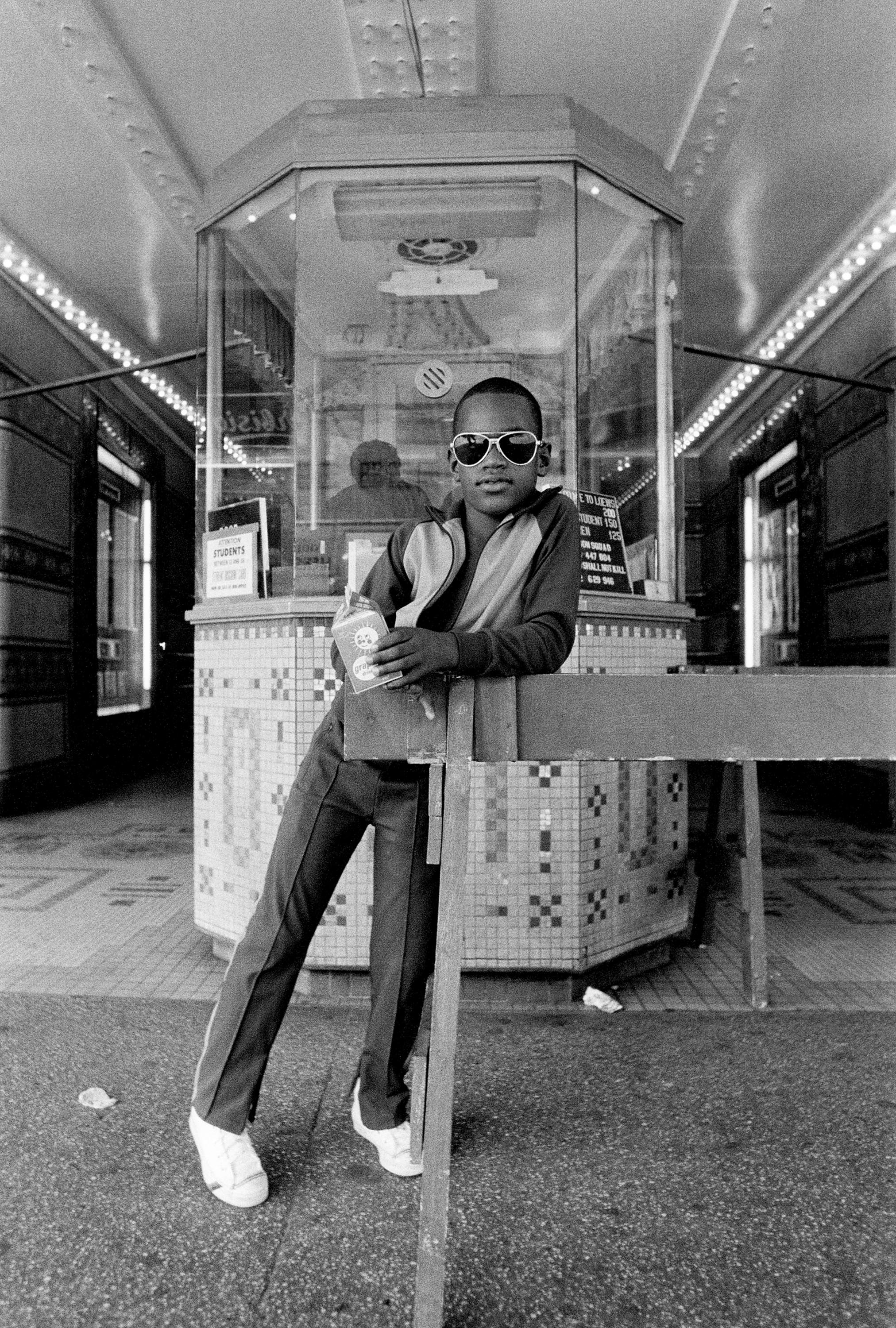 A black and white photograph of a person leaning against a counter inside a transit station, wearing sunglasses, a jacket, and pants, with a glass display case behind them.