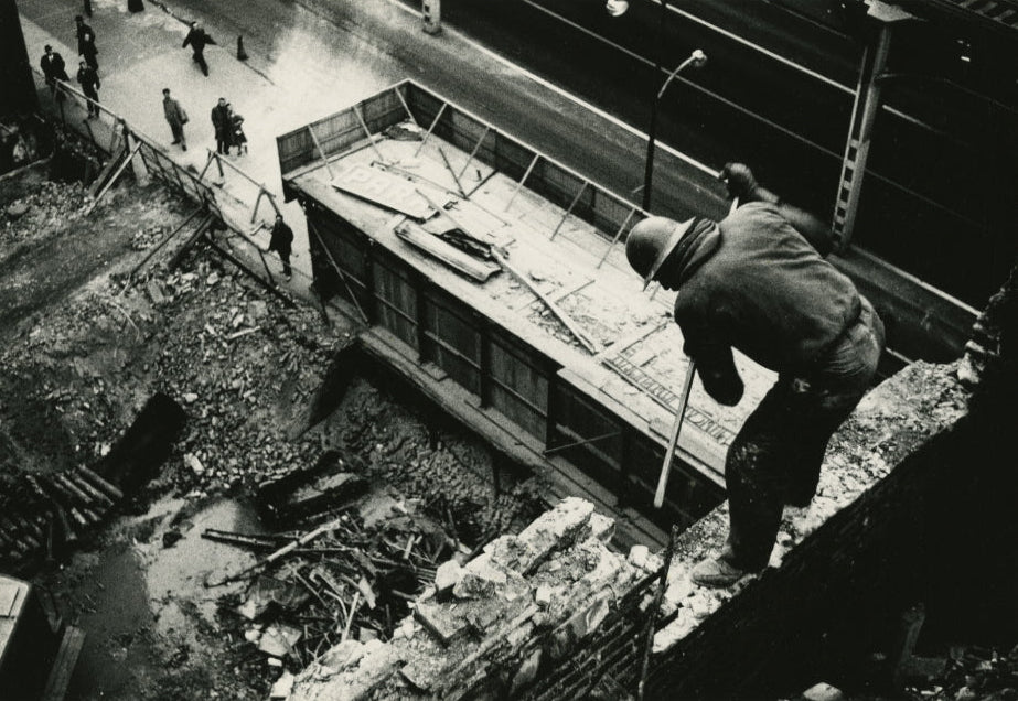 Black and white documentary-style photograph showing a worker in a hard hat standing on a crumbling brick wall above a debris-filled trench, with pedestrians and a street visible in the background.