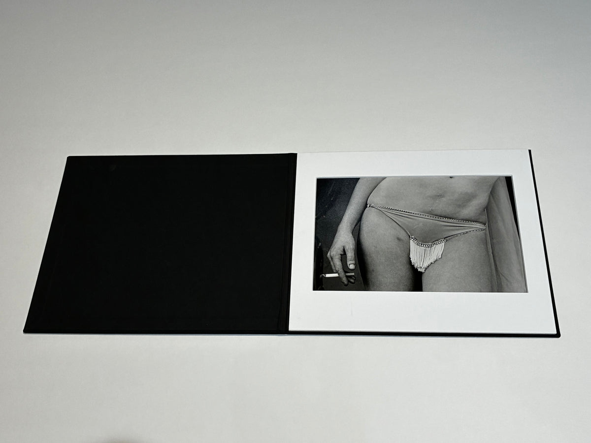 A black hardcover art book or photo album shown open on a light gray background, with a black-and-white photograph of a person wearing minimal underwear on the right page.